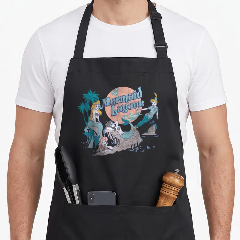 Petter distressed apron - Cartoon mermaids and boy around text Mermaid Lagoon over pink sun — shown on a black cotton canvas apron against a white background