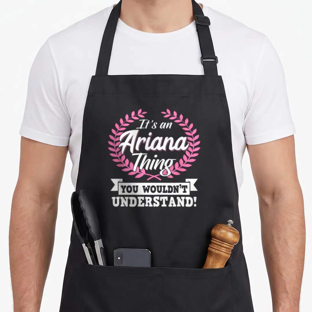 Ariana today smile apron - White text It's an Ariana Thing in pink laurel wreath above white ribbon banner — shown on a black cotton canvas apron against a white background
