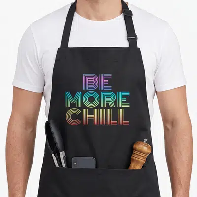Chill rainbow apron - Stacked BE MORE CHILL text in purple blue green yellow red retro stripes — shown on a black cotton canvas apron against a white background