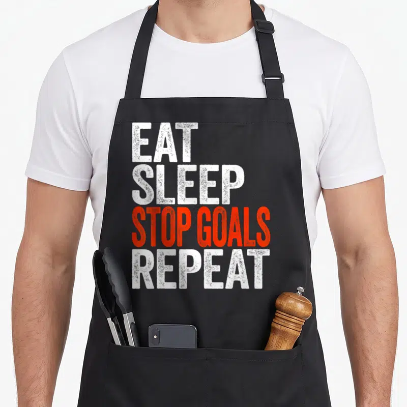 Eat goalkeeper apron - Stacked distressed text EAT SLEEP STOP GOALS REPEAT in white and red — shown on a black cotton canvas apron against a white background
