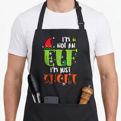 Xmas elf apron - Stacked text I'M NOT AN ELF I'M JUST SHORT with elf hat shoe and snowflakes — shown on a black cotton canvas apron against a white background