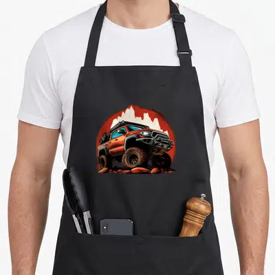 Tacoma crawler apron - Cartoon orange off-road truck climbing rocks inside orange circle with white mountain silhouettes — shown on a black cotton canvas apron against a white background