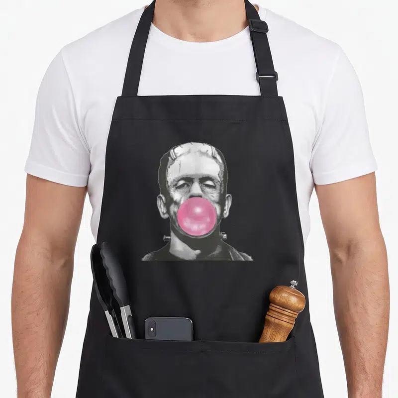 Frankenstein bubble gum apron - Grayscale Frankenstein head with pink bubblegum bubble covering mouth on black apron — shown on a black cotton canvas apron against a white background