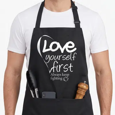 Yourself lettering quotes apron - White heart outline wrapping stacked text next to a small fist icon — shown on a black cotton canvas apron against a white background