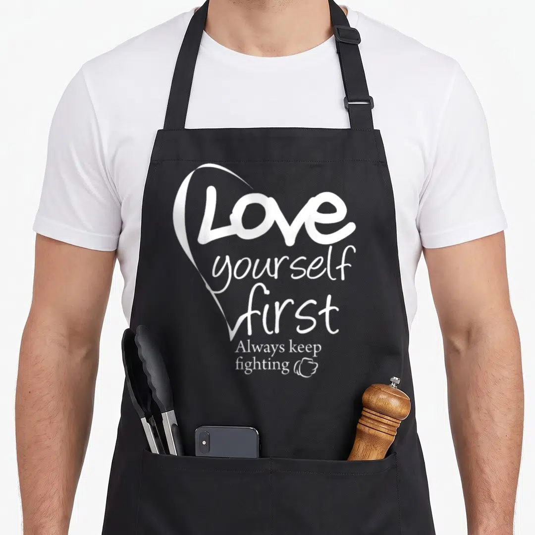 Yourself lettering quotes apron - White heart outline wrapping stacked text next to a small fist icon — shown on a black cotton canvas apron against a white background