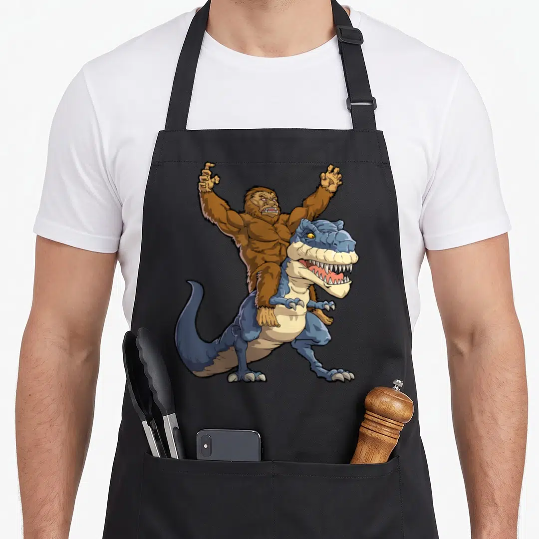 Bigfoot dinosaur apron - Brown gorilla riding blue t-rex cartoon illustration with beige belly details — shown on a black cotton canvas apron against a white background