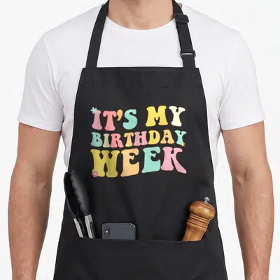 Week born day apron - IT'S MY BIRTHDAY WEEK text in pink blue yellow orange bubbly font with small flowers — shown on a black cotton canvas apron against a white background