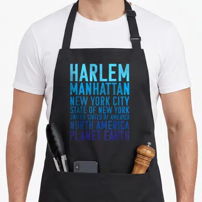 Planet new york apron - Stacked text reading Harlem Manhattan New York City State USA North America Planet Earth — shown on a black cotton canvas apron against a white background
