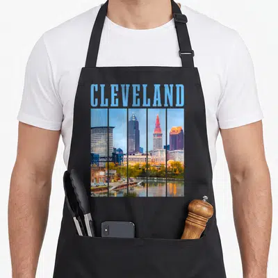 Cleveland skyline pride apron - Retro Cleveland skyline in five vertical panels with light blue CLEVELAND text above — shown on a black cotton canvas apron against a white background