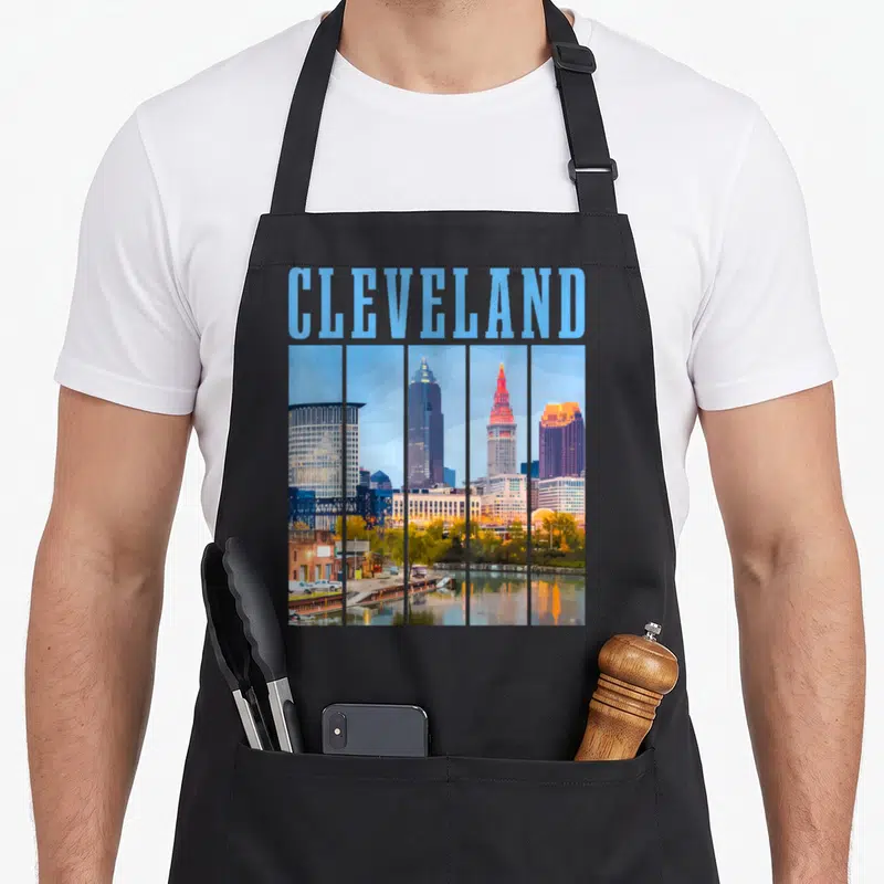 Cleveland skyline pride apron - Retro Cleveland skyline in five vertical panels with light blue CLEVELAND text above — shown on a black cotton canvas apron against a white background