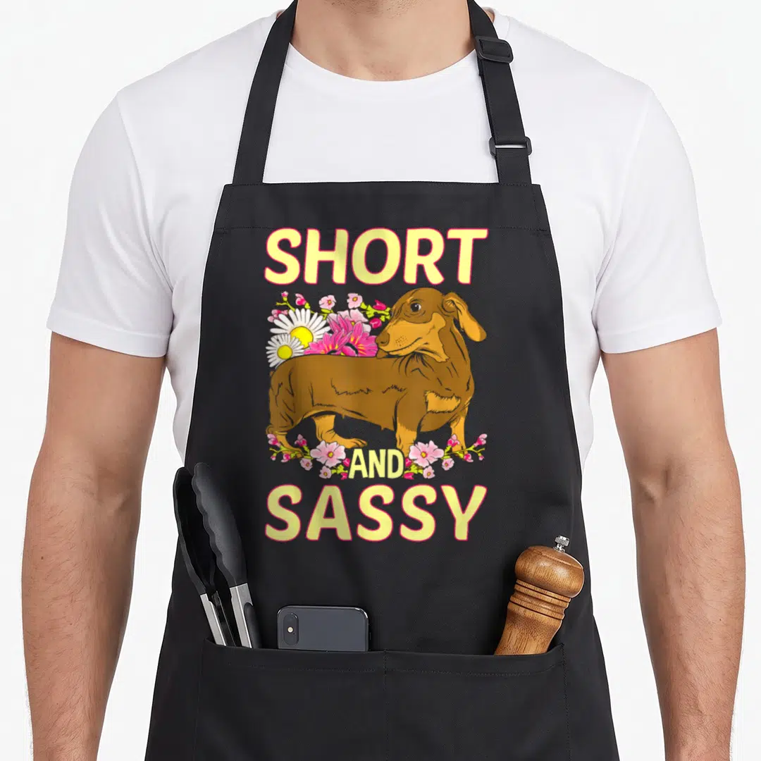 Sassy dachshund apron - Cartoon brown dachshund with Short and Sassy text and pink yellow flowers — shown on a black cotton canvas apron against a white background