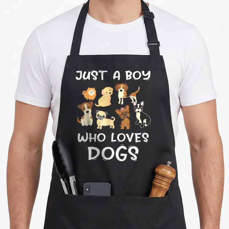Cute puppy dog apron - Seven cartoon dogs in orange and brown flanked by white text reading JUST A BOY WHO LOVES DOGS — shown on a black cotton canvas apron against a white background