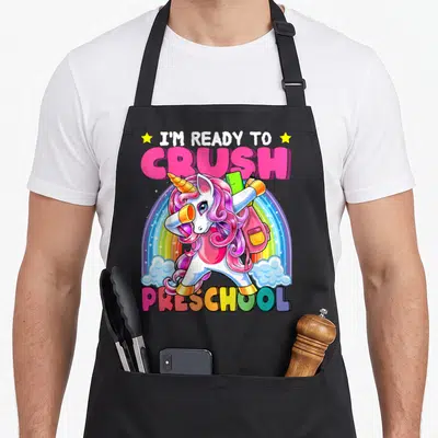 Crush preschool apron - Dabbing unicorn with backpack in front of rainbow and clouds under text I'm ready to crush — shown on a black cotton canvas apron against a white background