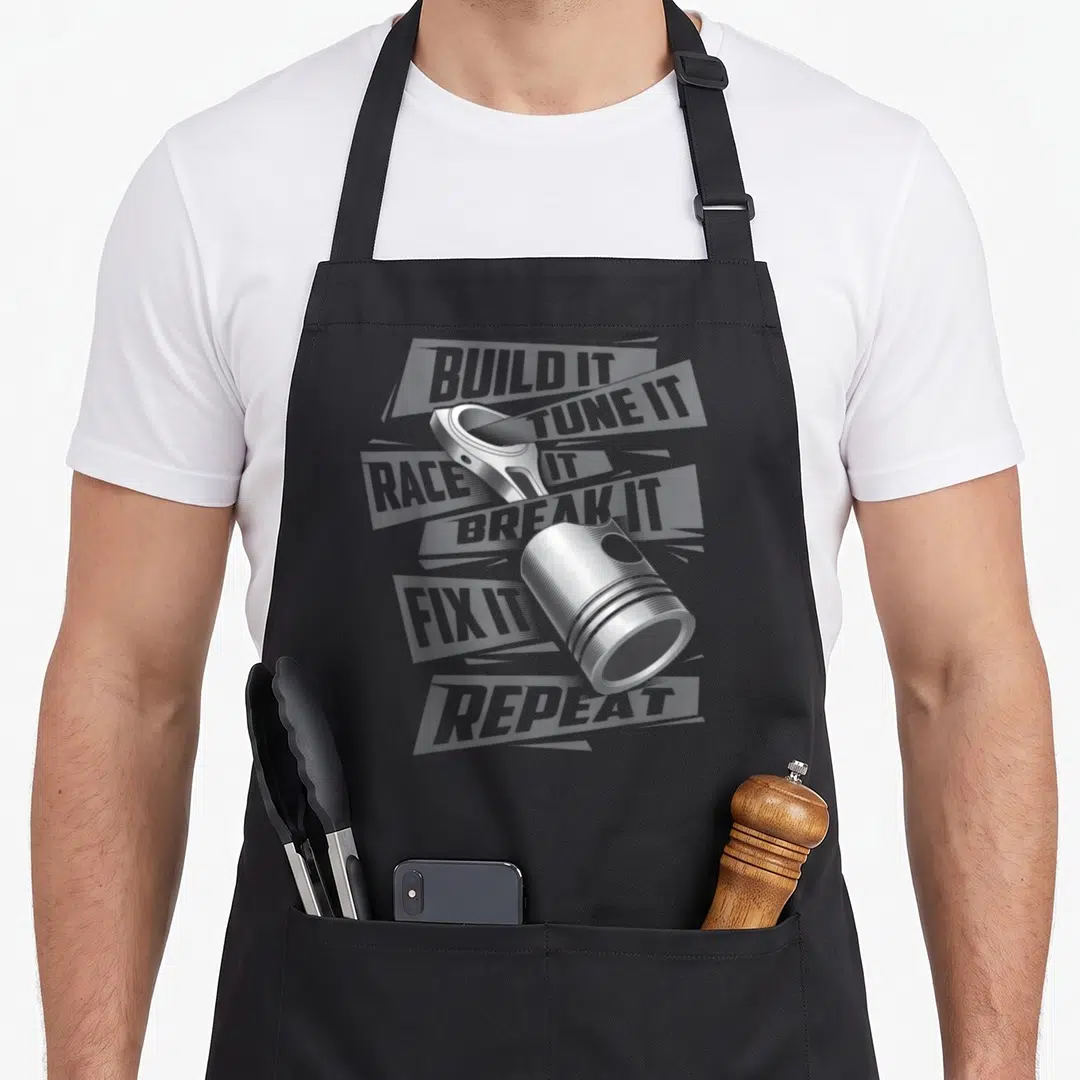 Break car apron - Silver piston graphic overlaid on grey banners with BUILD IT TUNE IT RACE IT BREAK IT FIX IT REPEAT — shown on a black cotton canvas apron against a white background