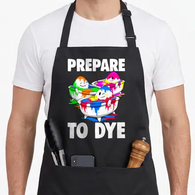 Tea easter apron - PREPARE TO DYE text above three cartoon eggs in bathtubs colored white blue green pink yellow — shown on a black cotton canvas apron against a white background