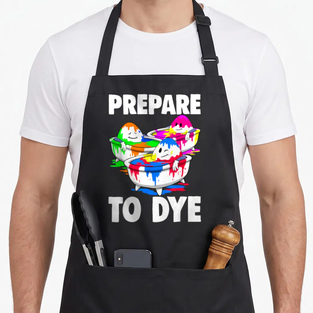 Tea easter apron - PREPARE TO DYE text above three cartoon eggs in bathtubs colored white blue green pink yellow — shown on a black cotton canvas apron against a white background