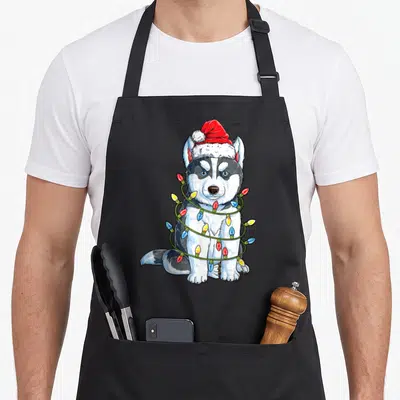 Santa husky apron - Cartoon husky wearing red Santa hat wrapped in green string lights with colored bulbs — shown on a black cotton canvas apron against a white background