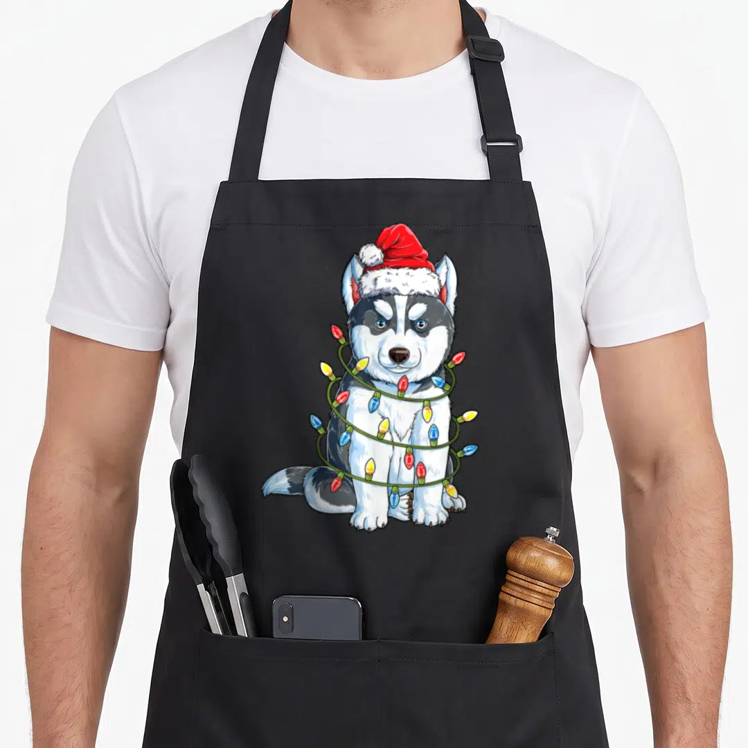Santa husky apron - Cartoon husky wearing red Santa hat wrapped in green string lights with colored bulbs — shown on a black cotton canvas apron against a white background