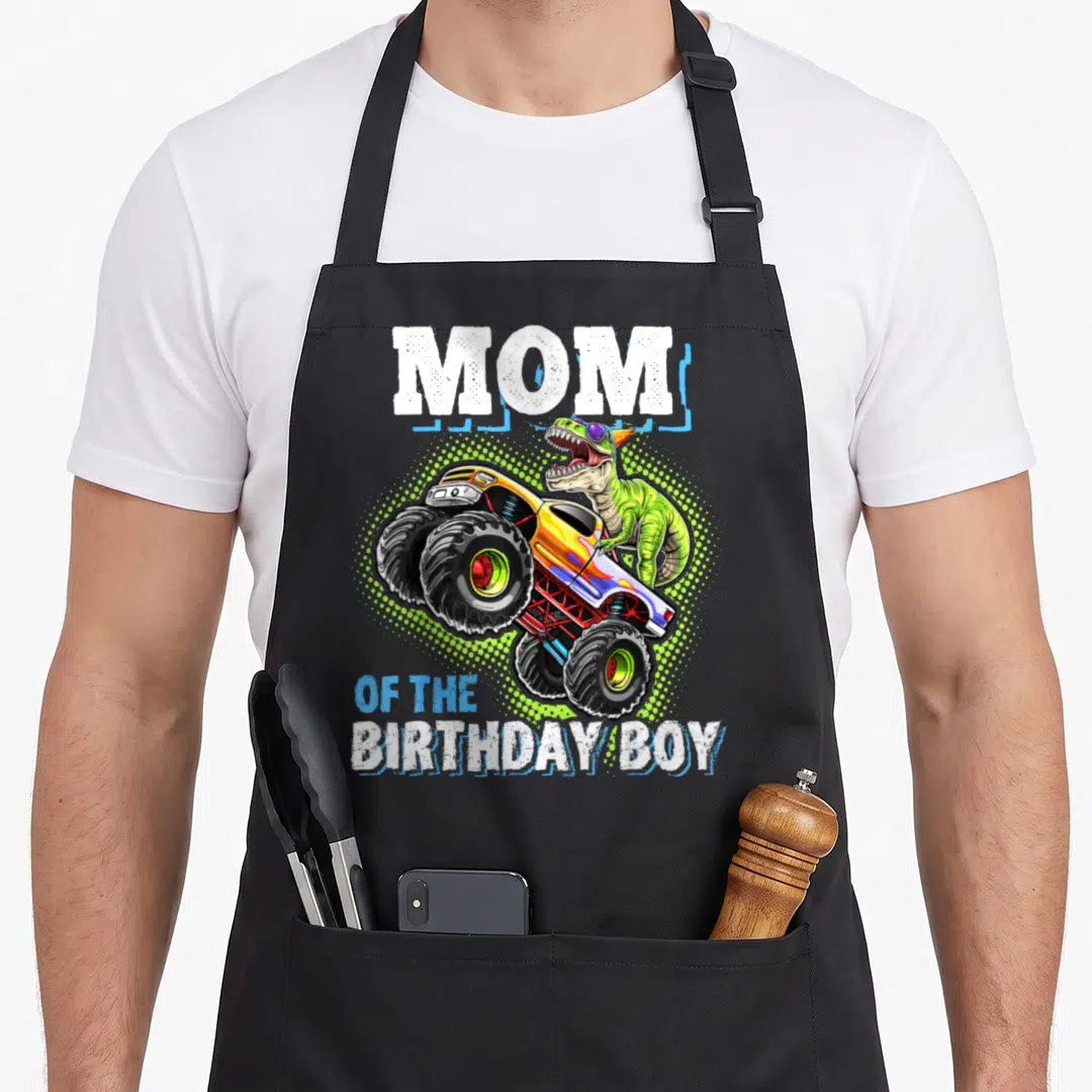 Birthday dinosaur truck apron - MOM OF THE BIRTHDAY BOY text with green dinosaur riding monster truck over halftone dots — shown on a black cotton canvas apron against a white background