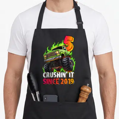 Monster 5th birthday truck apron - Green monster truck with flames and crushin it since 2019 text — shown on a black cotton canvas apron against a white background