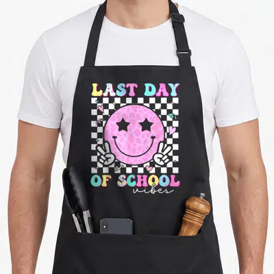 Student groovy teacher apron - Pink smiley face with star eyes and peace hands on checkerboard with last day of school — shown on a black cotton canvas apron against a white background