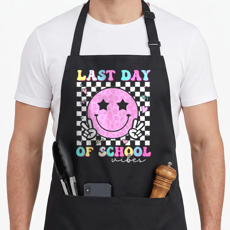 Student groovy teacher apron - Pink smiley face with star eyes and peace hands on checkerboard with last day of school — shown on a black cotton canvas apron against a white background
