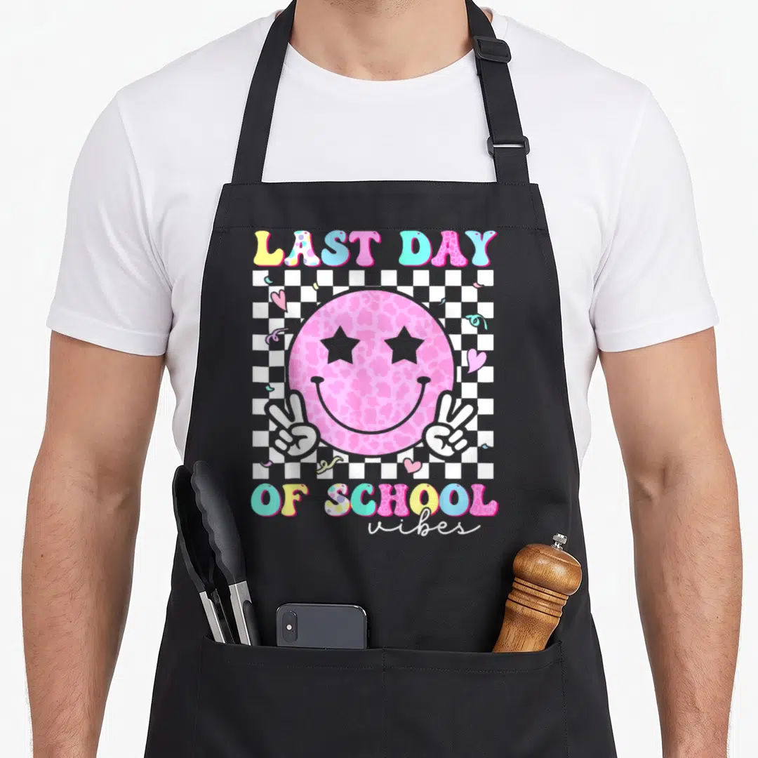 Student groovy teacher apron - Pink smiley face with star eyes and peace hands on checkerboard with last day of school — shown on a black cotton canvas apron against a white background