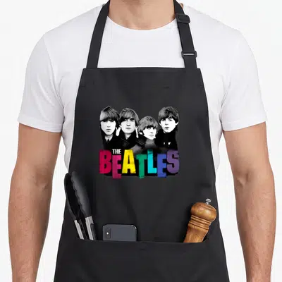 Vintage colorful aprons - Four men portraits with distressed colorful THE BEATLES text in pink yellow green blue purple — shown on a black cotton canvas apron against a white background