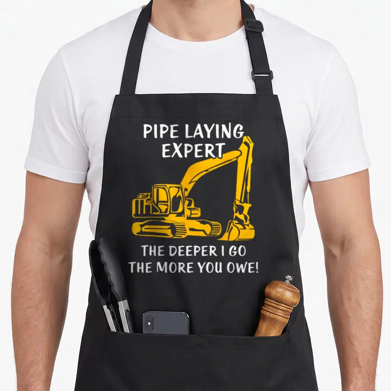 Pipe heavy canvas aprons - Yellow excavator graphic with white text pipe laying expert and deeper I go more you owe — shown on a black cotton canvas apron against a white background