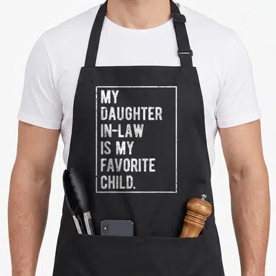 Cool daughter father day apron - White stacked text MY DAUGHTER IN-LAW IS MY FAVORITE CHILD inside distressed white — shown on a black cotton canvas apron against a white background