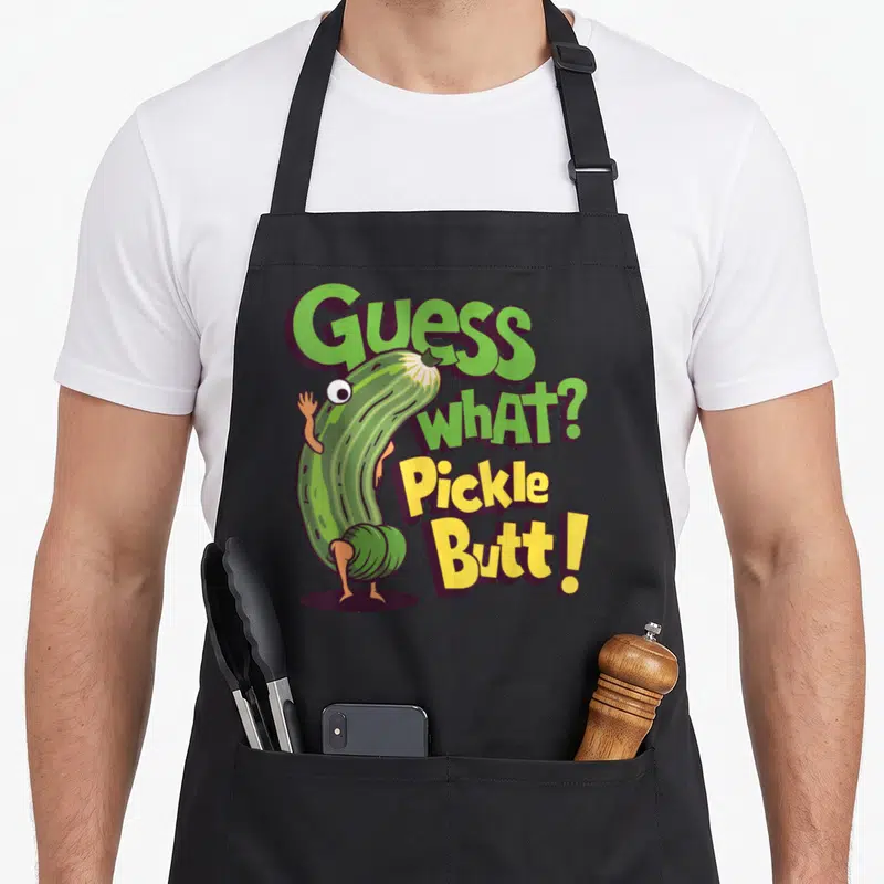 Guess meme apron - Cartoon pickle bending over with text Guess What Pickle Butt in green and yellow — shown on a black cotton canvas apron against a white background