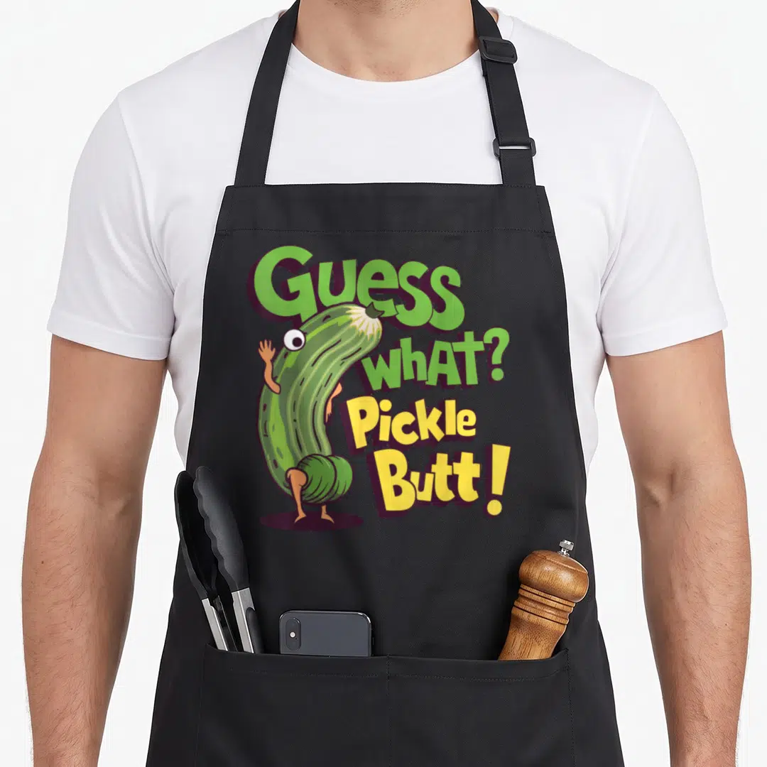 Guess meme apron - Cartoon pickle bending over with text Guess What Pickle Butt in green and yellow — shown on a black cotton canvas apron against a white background
