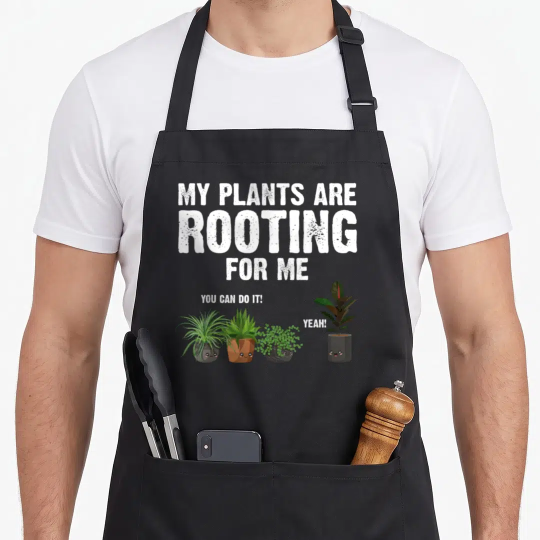 Funny gardening gardener apron - Four cartoon potted plants with faces and speech bubbles under rooting pun text — shown on a black cotton canvas apron against a white background