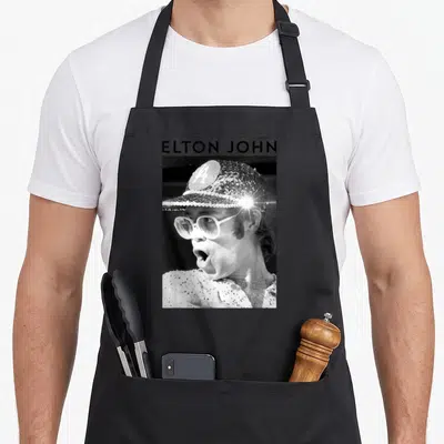 Elton sequin apron - ELTON JOHN text above black and white photo of man in sequined cap and glasses — shown on a black cotton canvas apron against a white background