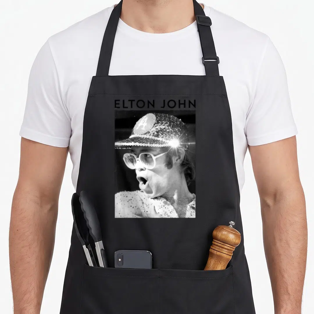 Elton sequin apron - ELTON JOHN text above black and white photo of man in sequined cap and glasses — shown on a black cotton canvas apron against a white background