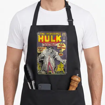 Incredible book apron - Vintage Hulk comic cover print with yellow title and speech bubbles asking man or monster — shown on a black cotton canvas apron against a white background