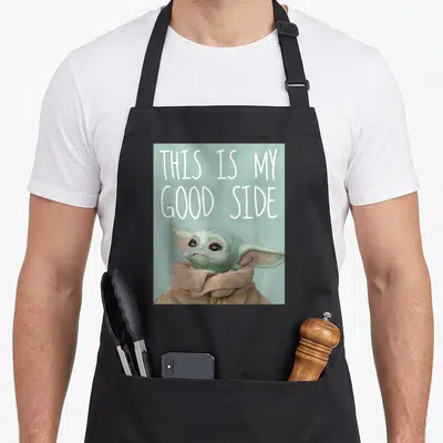 Funny food novelty apron - Grogu character with THIS IS MY GOOD SIDE text on light blue background — shown on a black cotton canvas apron against a white background