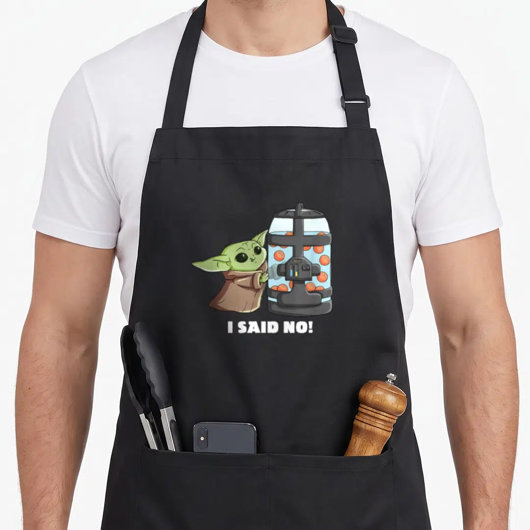 Frog egg apron - Grogu holding orange egg container with I said no text below — shown on a black cotton canvas apron against a white background