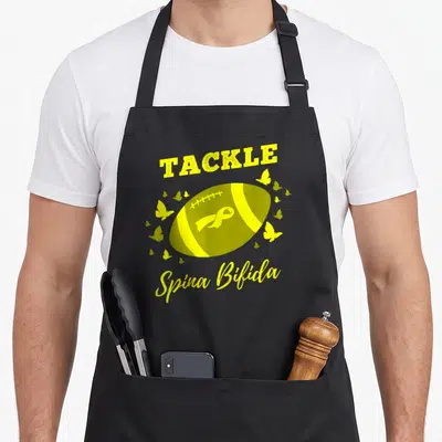 Tackle spina bifida awareness apron - Yellow football with ribbon and butterflies surrounded by text TACKLE Spina Bifida — shown on a black cotton canvas apron against a white background