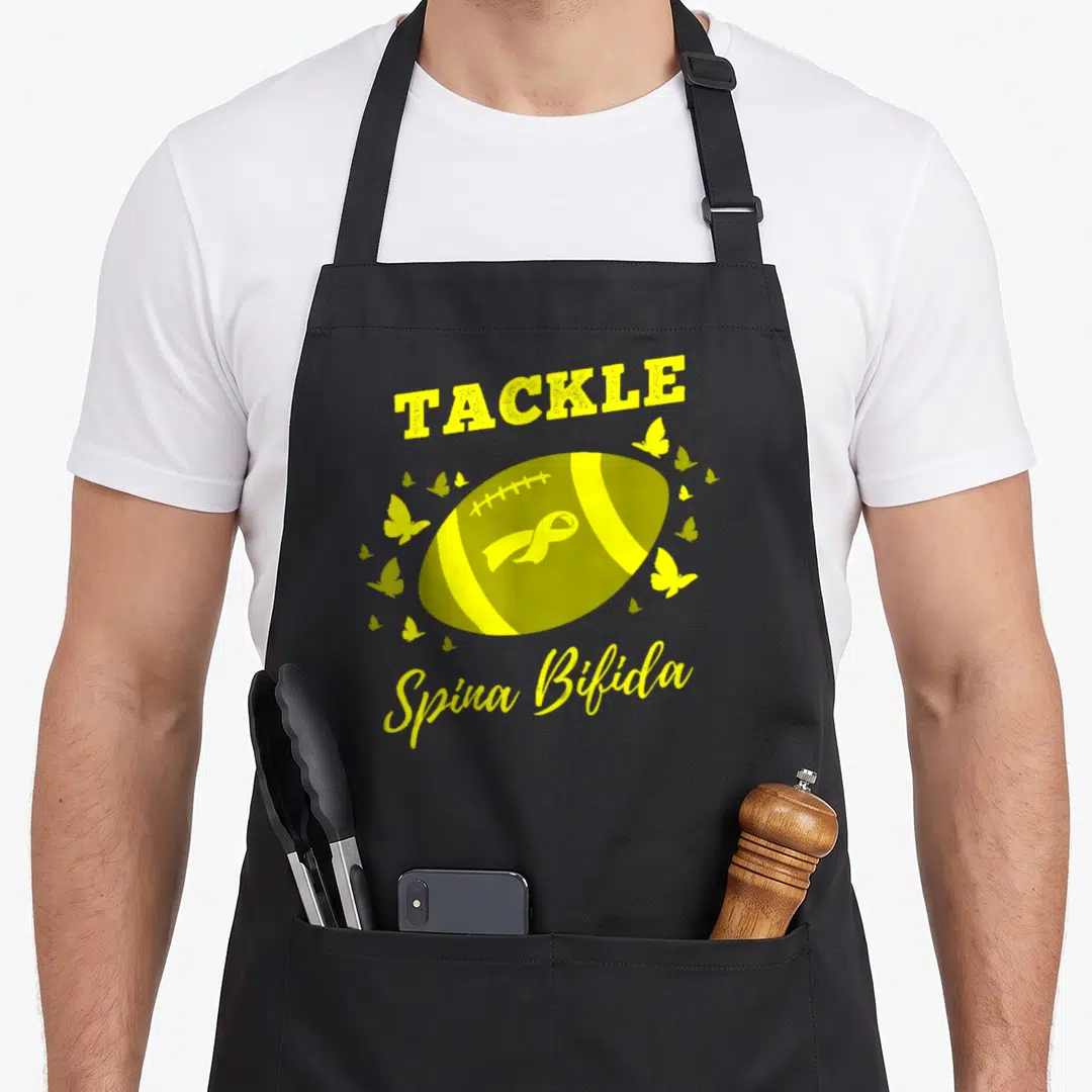 Tackle spina bifida awareness apron - Yellow football with ribbon and butterflies surrounded by text TACKLE Spina Bifida — shown on a black cotton canvas apron against a white background