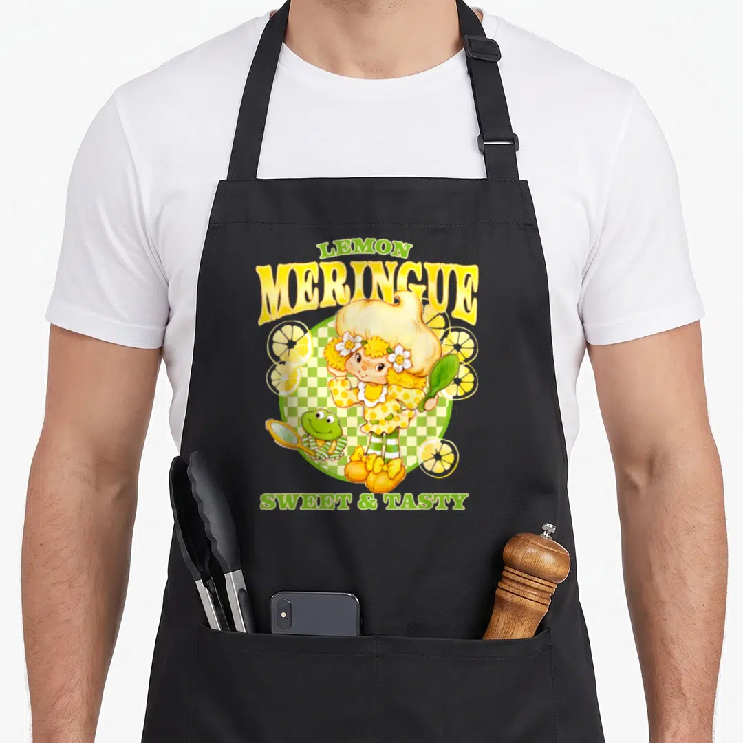 Strawberry lemon apron - Cartoon girl standing before checkered circle with frog, lemon slices, and meringue text — shown on a black cotton canvas apron against a white background