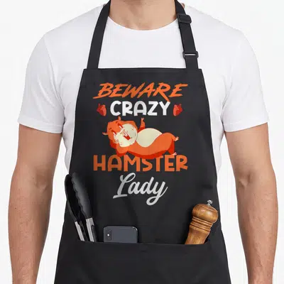 Beware crazy hamster man apron - Cartoon orange hamster with BEWARE CRAZY HAMSTER Lady text and two acorns — shown on a black cotton canvas apron against a white background