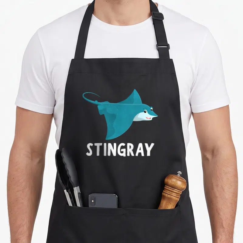 Funny stingray apron or apron - Blue cartoon stingray illustration above white STINGRAY text — shown on a black cotton canvas apron against a white background