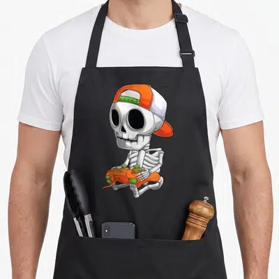 Skeleton video halloween apron - Cartoon skeleton sitting with orange cap and game controller in white, orange — shown on a black cotton canvas apron against a white background