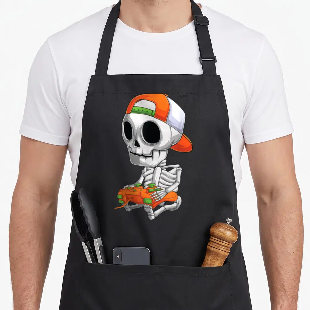 Skeleton video halloween apron - Cartoon skeleton sitting with orange cap and game controller in white, orange — shown on a black cotton canvas apron against a white background