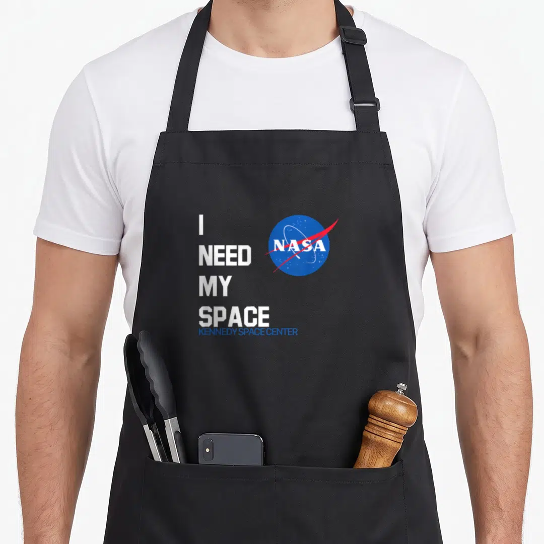 Kennedy nasa apron - I NEED MY SPACE text next to blue red NASA meatball logo — shown on a black cotton canvas apron against a white background