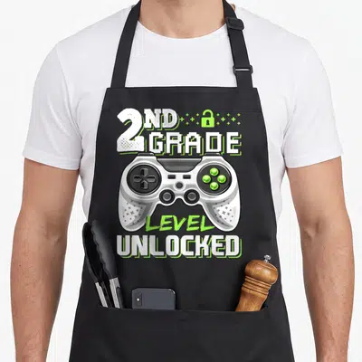 2nd video game apron - Controller graphic with 2ND GRADE LEVEL UNLOCKED text in white lime green and grey — shown on a black cotton canvas apron against a white background