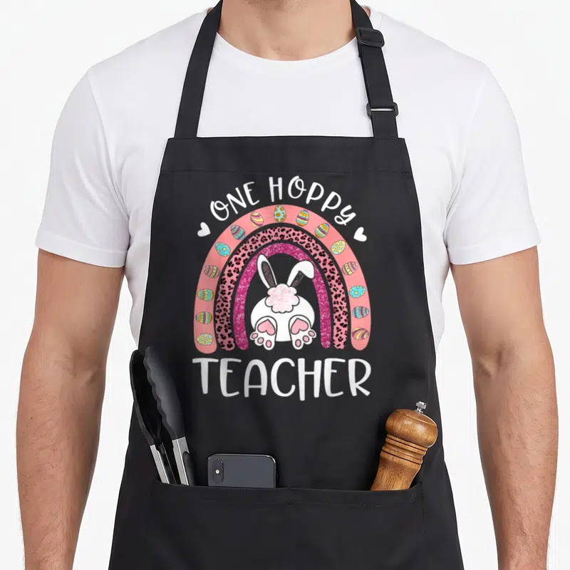 Hoppy teacher apron - ONE HOPPY TEACHER text over rainbow with bunny back, leopard stripe, and Easter eggs — shown on a black cotton canvas apron against a white background