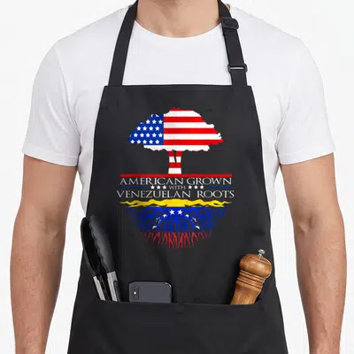 Root grown american flag apron - Tree with American flag canopy and Venezuelan roots displaying AMERICAN GROWN — shown on a black cotton canvas apron against a white background
