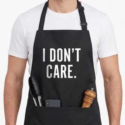 Funny care sarcastic apron - White I DON'T CARE text stacked on two lines against a dark background — shown on a black cotton canvas apron against a white background